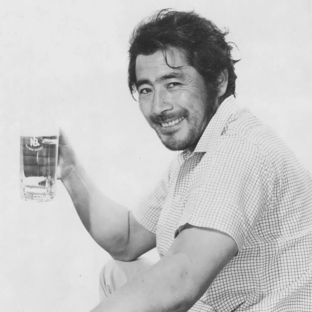 Japanese actor Toshiro Mifune photographed against a white background with a heartthrob smile and holding what could be a glass of beer, but it's hard to tell because the photo is black and white