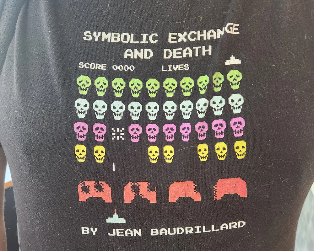 a t-shirt with invaders-style screenshot but the aliens are skulls. the text reads: "SYMBOLIC EXCHANGE AND DEATH; BY JEAN BAUDRILLARD"