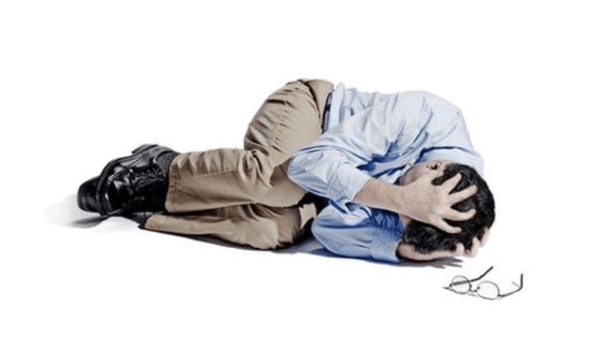 Stock photo of a person in business-casual clothes curled up in the fetal position on the floor of a white void gripping their head.