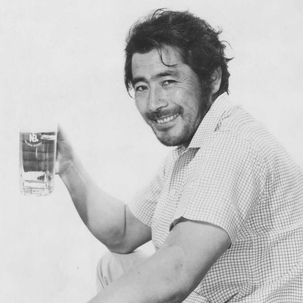 Japanese actor Toshiro Mifune photographed against a white background with a heartthrob smile and holding what could be a glass of beer, but it's hard to tell because the photo is black and white