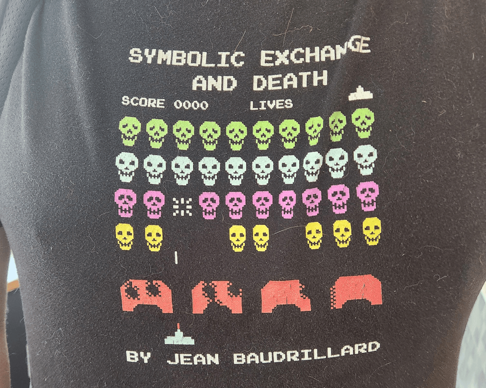 a t-shirt with invaders-style screenshot but the aliens are skulls. the text reads: "SYMBOLIC EXCHANGE AND DEATH; BY JEAN BAUDRILLARD"